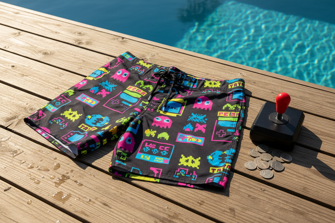 Retro gaming boardshorts with pixel art arcade design displayed poolside with vintage joystick and quarters — Caster Coast